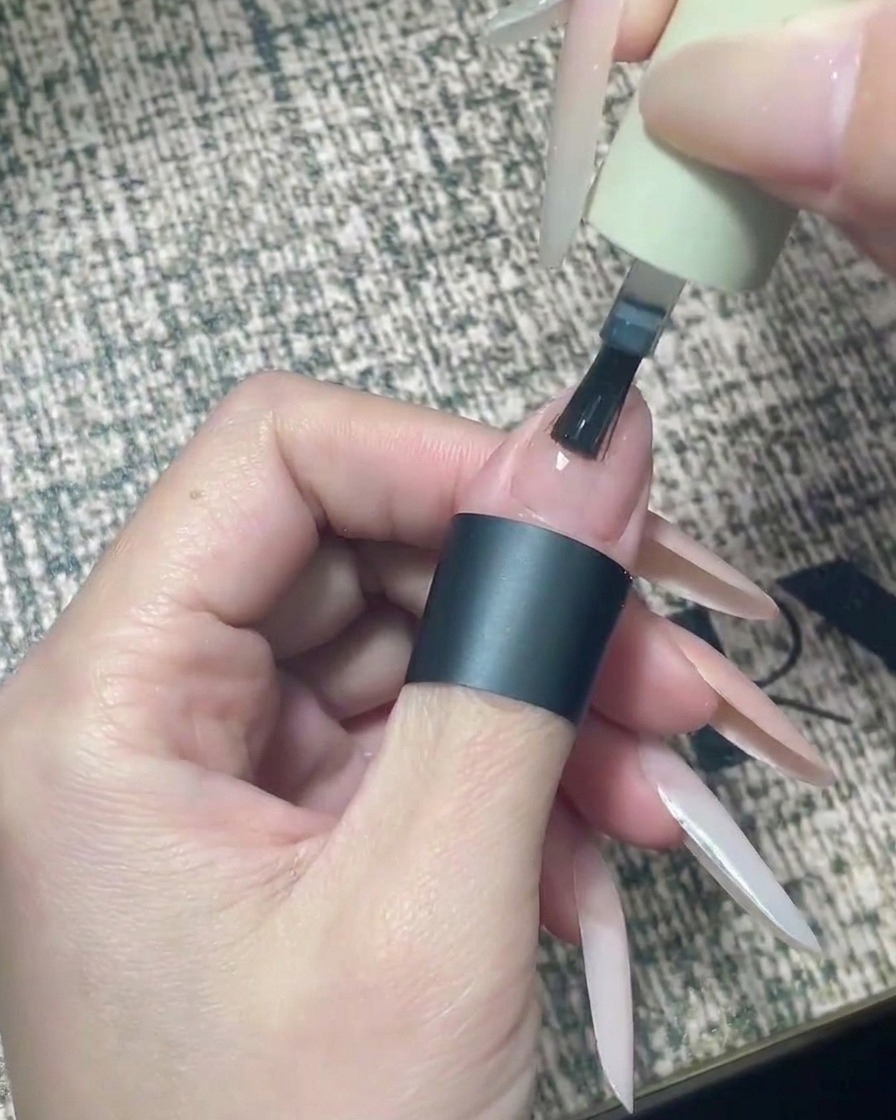 Un pincel aplicando top coat transparente con precisión en la mitad superior de una uña color nude.
