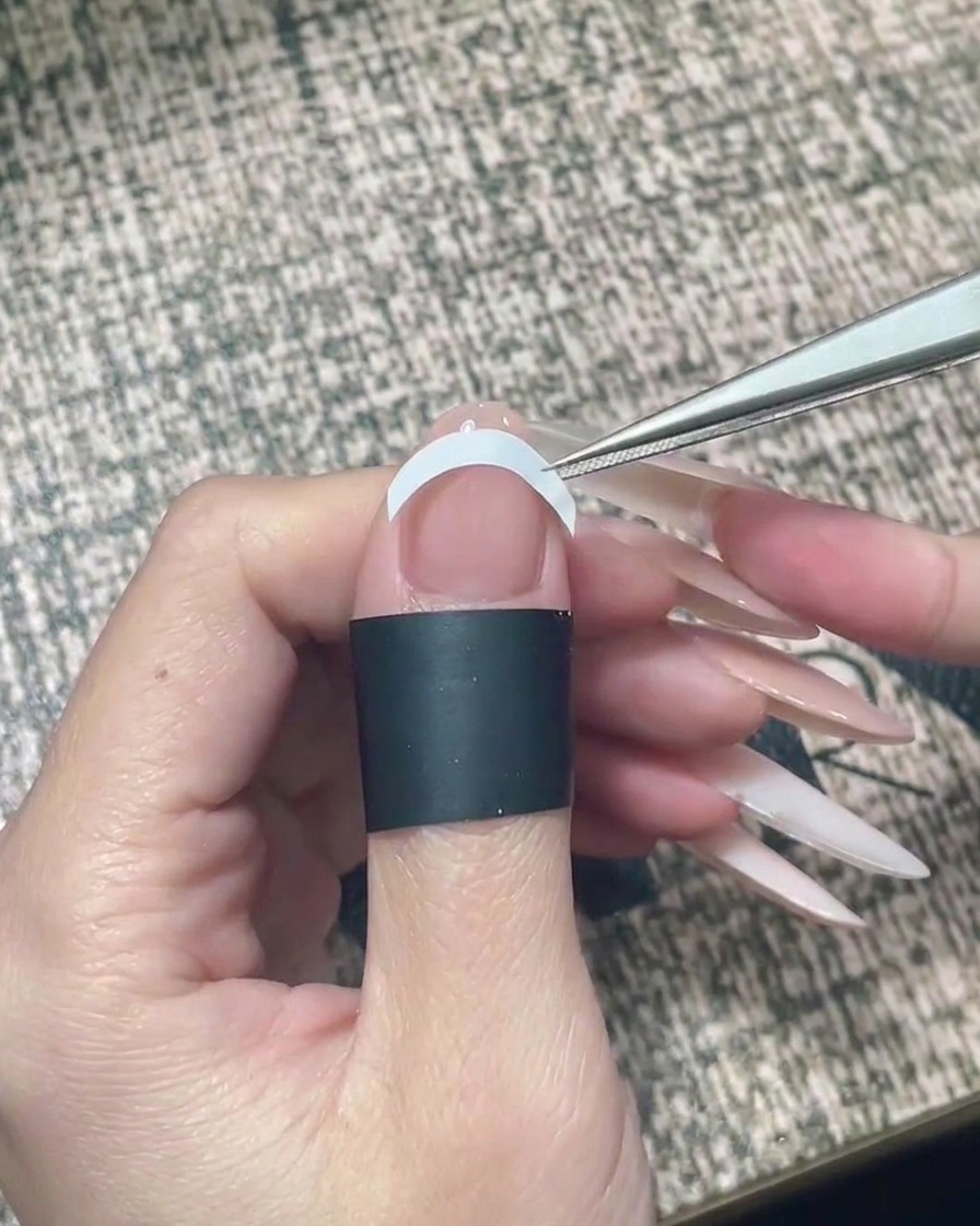 Unas pinzas colocando una pegatina blanca curva como guía de manicura francesa horizontalmente en el medio de la uña.
