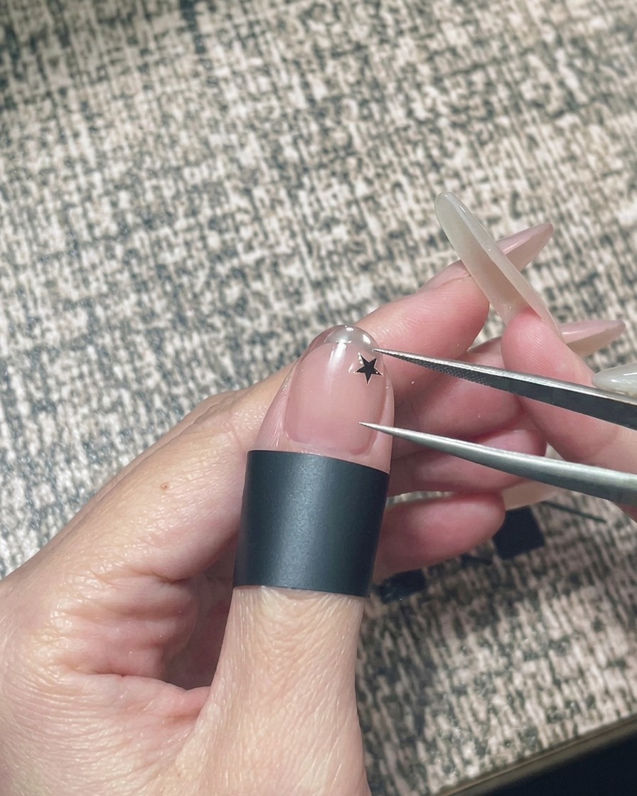Una mano sosteniendo unas pinzas para colocar una pequeña pegatina de estrella negra sobre una uña de gel nude.