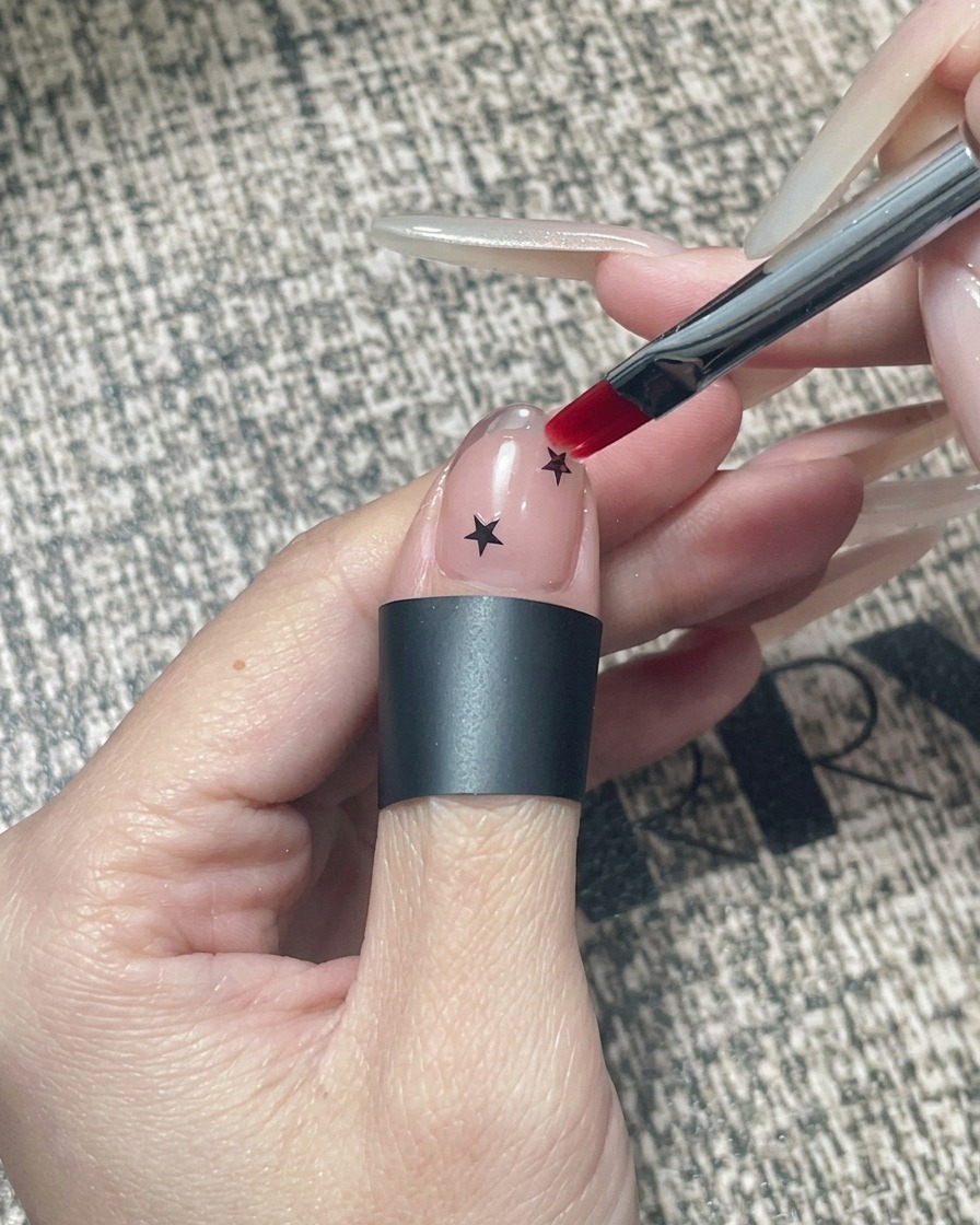 Un pincel cuadrado para gel aplicando gel transparente para encapsular sobre una uña decorada con estrellas negras y una punta cromada.