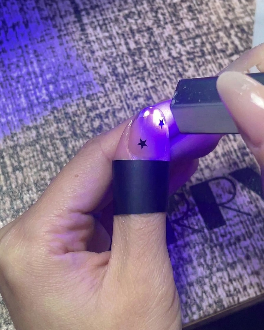 Primer plano de una mano utilizando una pequeña linterna UV de mano para dar un curado rápido a una uña decorada con pegatinas de estrellas negras.