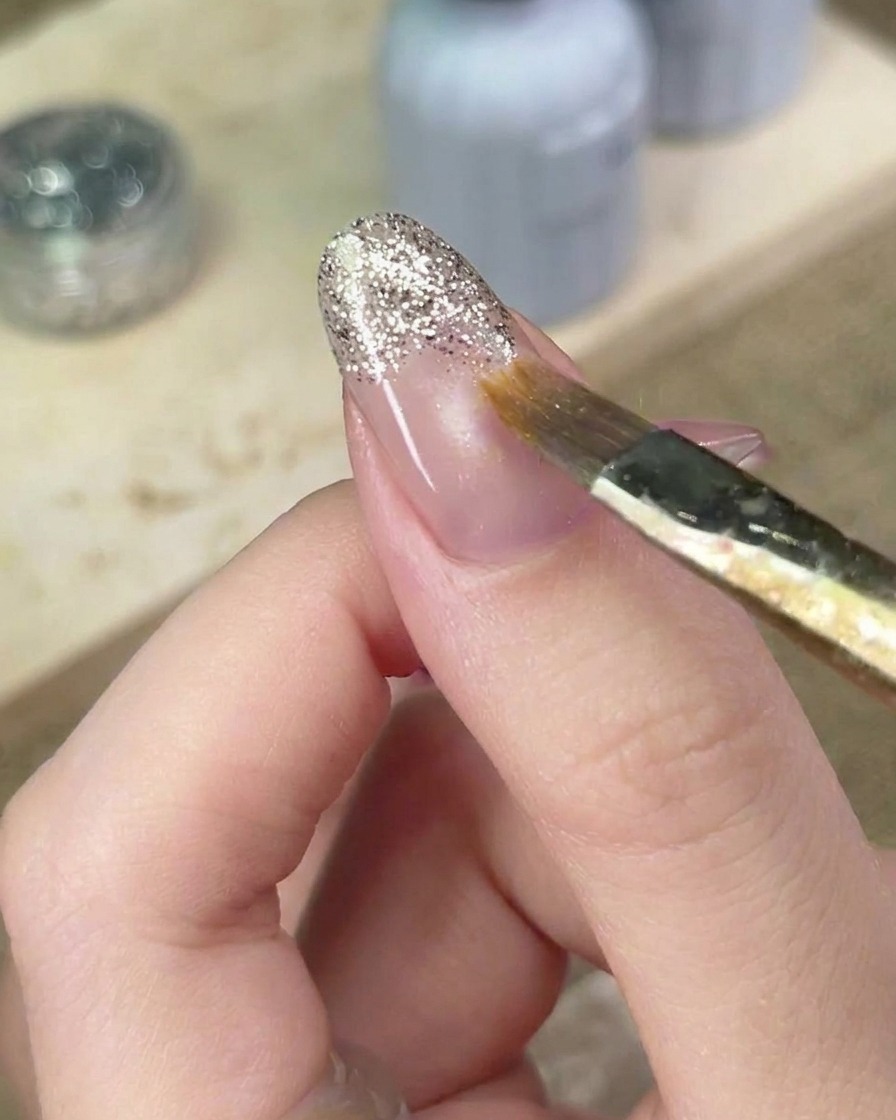 Un pincel de detalle difuminando con cuidado los bordes del gel de purpurina plateada en la uña para perfeccionar el degradado.