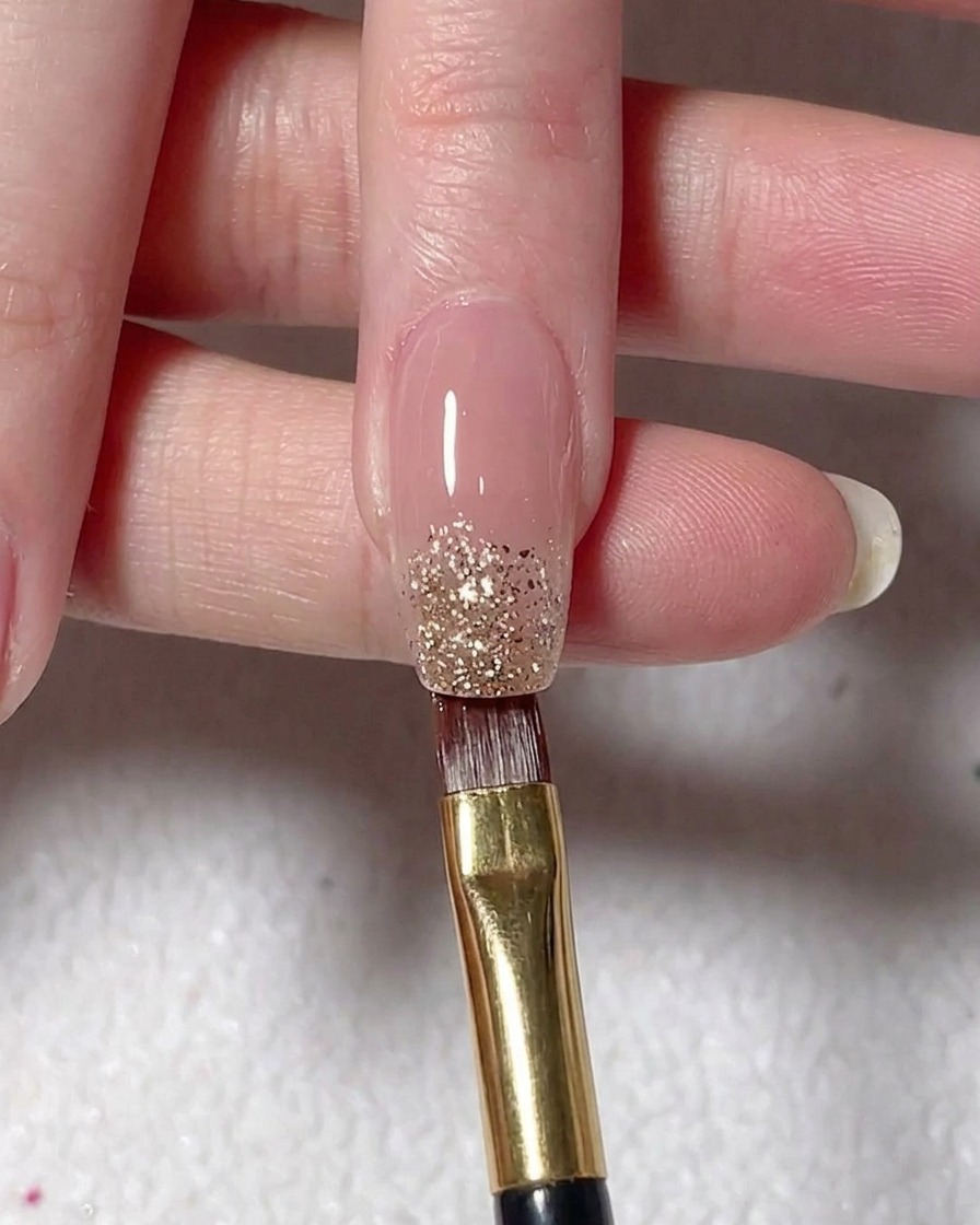 Un pincel plano depositando gel con glitter plateado en la punta de una uña pintada de color nude.