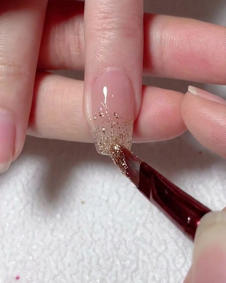 Un pincel fino difuminando el glitter plateado hacia arriba desde la punta de la uña para crear un efecto ombré.