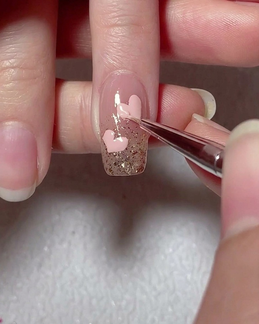 Un pincel liner refinando los bordes de un corazón de gel rosa sobre una uña decorada con glitter.