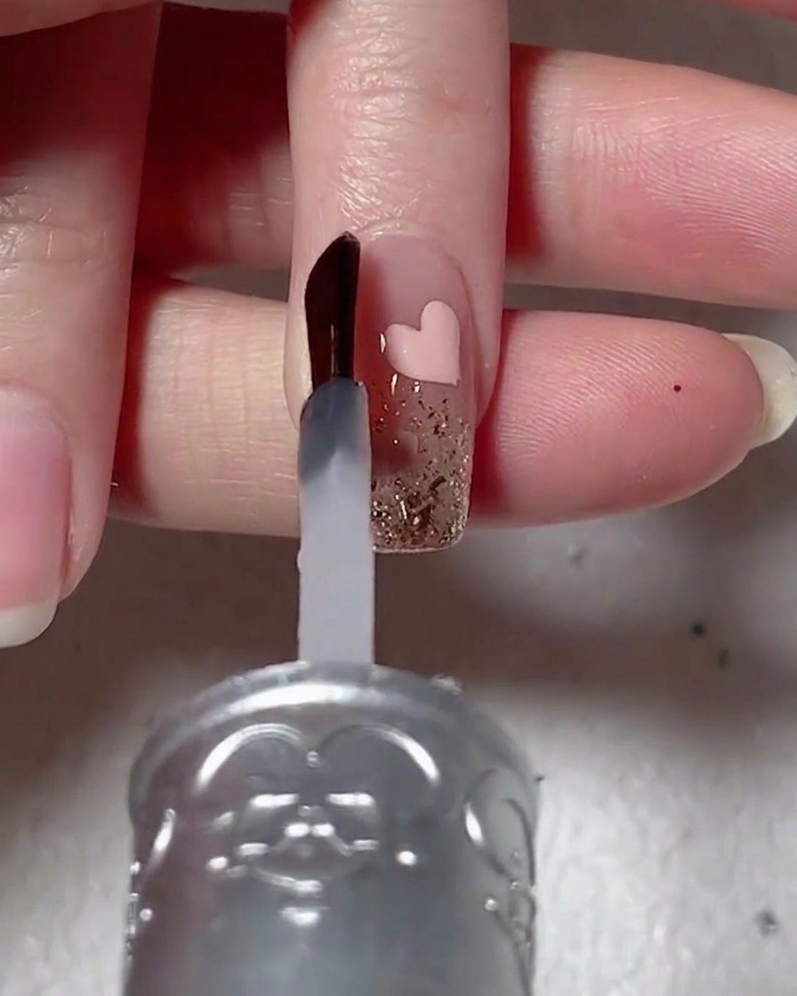 Un pincel de uñas aplicando un gel de refuerzo transparente y viscoso sobre una uña decorada con glitter y un corazón rosa.