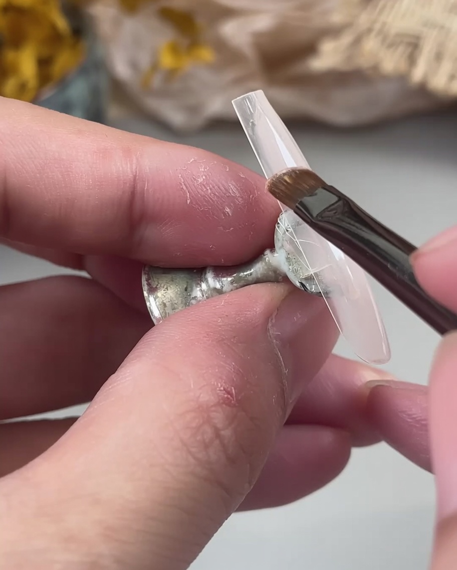 Un pincel aplicando una base coat de gel nude lechosa sobre un tip de uña transparente sostenido por dedos.