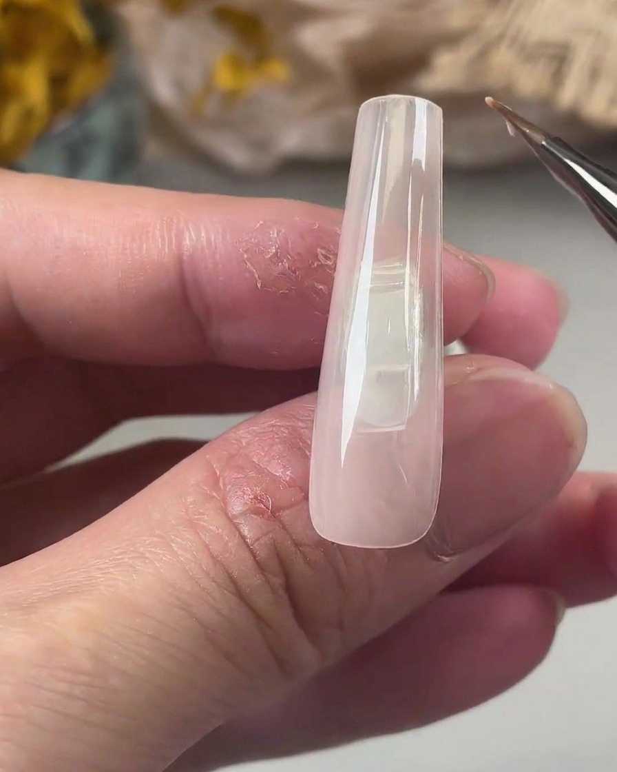 Un pincel fino de gel aplicando una capa suave de base coat transparente sobre una uña cuadrada color nude.