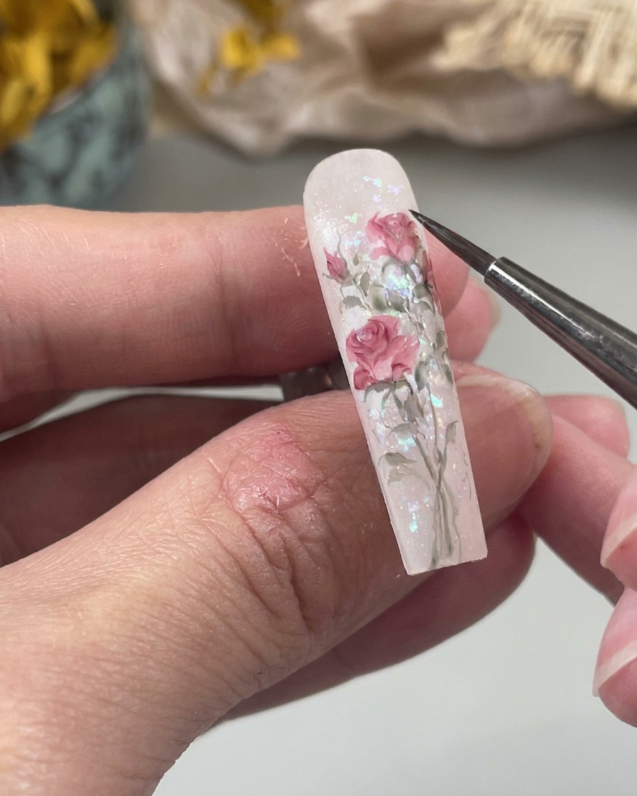 Primer plano de un pincel fino pintando una pequeña forma suave de gota de agua rosa sobre una uña como parte del diseño floral.