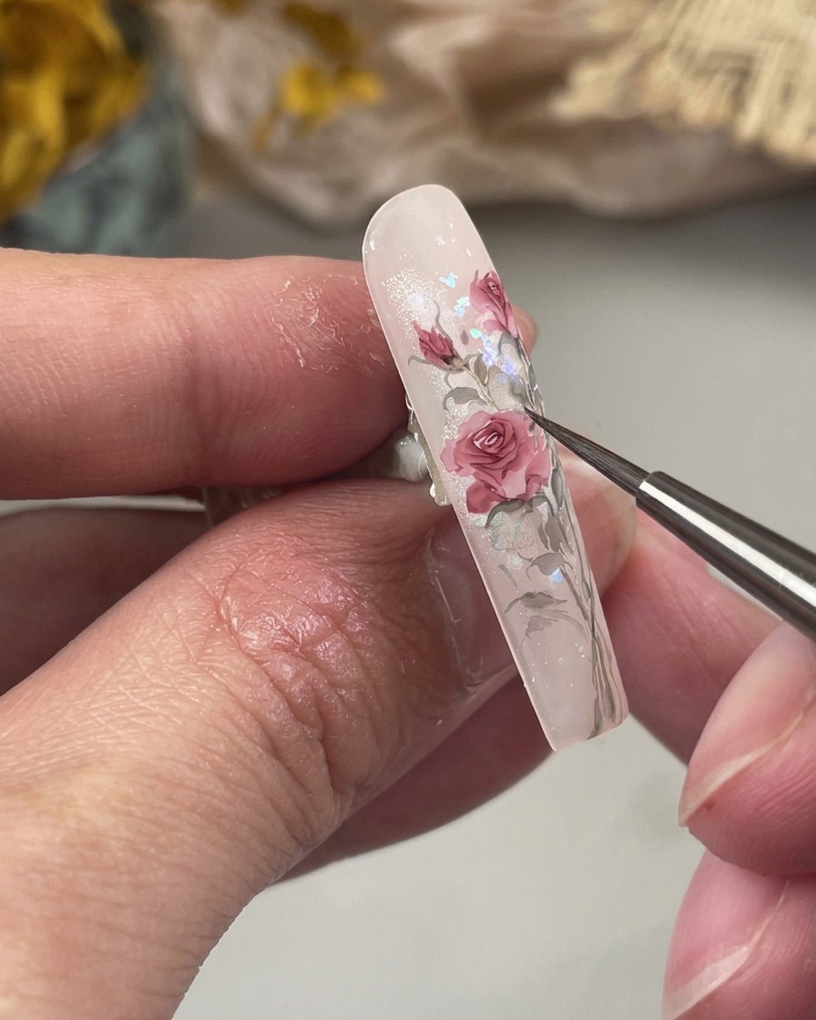 Una toma de primer plano mostrando un pincel fino pintando pequeñas hojas verdes afiladas en la base del arte floral de la uña.
