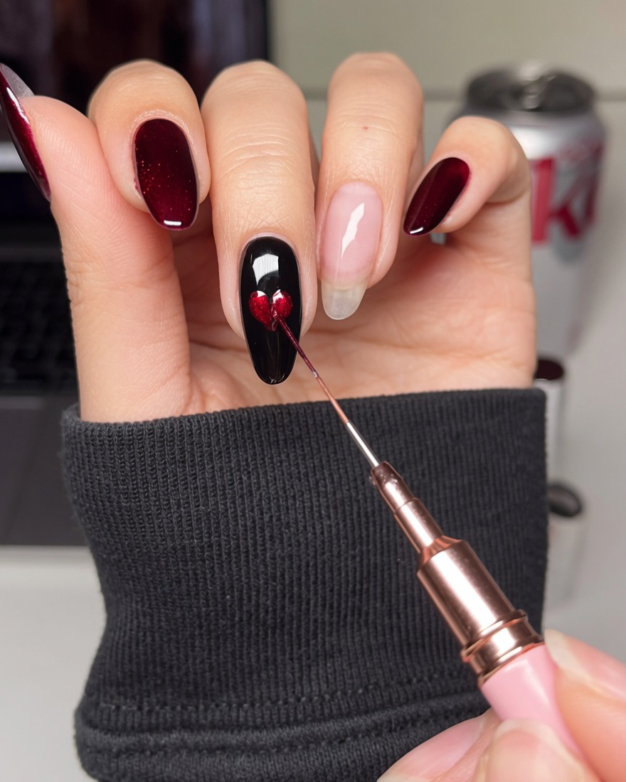 Un pincel de detalle fino rellenando el centro de una plantilla de corazón en una uña negra con esmalte en gel rojo.