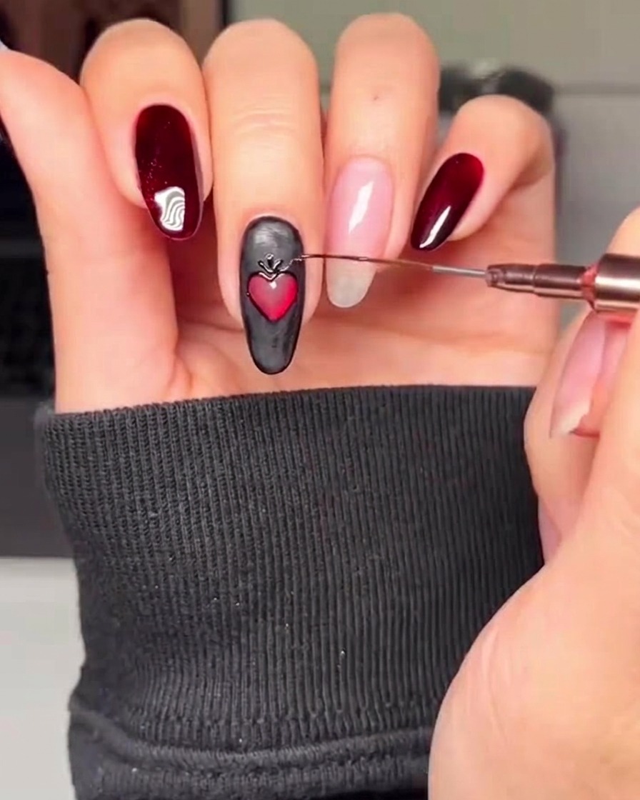 Primer plano de una uña negra con un corazón rojo, con un pincel fino añadiendo acentos decorativos dorados alrededor del perímetro del corazón.