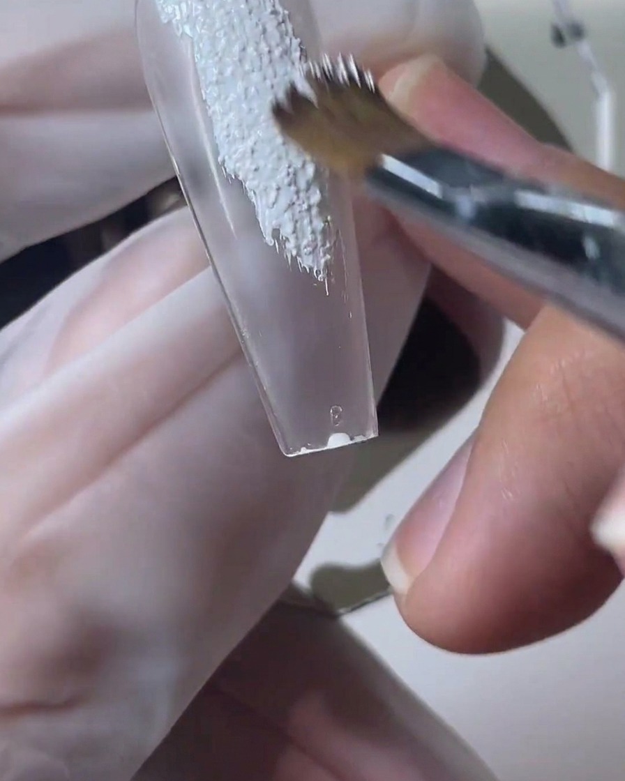Un pincel de uñas aplicando gel de arena texturizado blanco sobre un tip transparente.