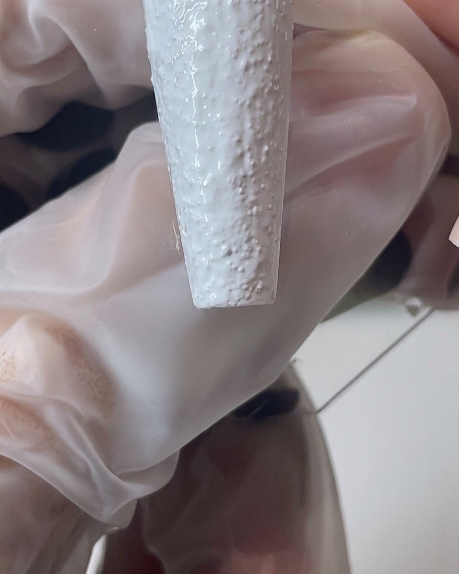 Primer plano de un pincel manipulando gel de arena blanco para crear un borde texturizado más grueso en la uña.
