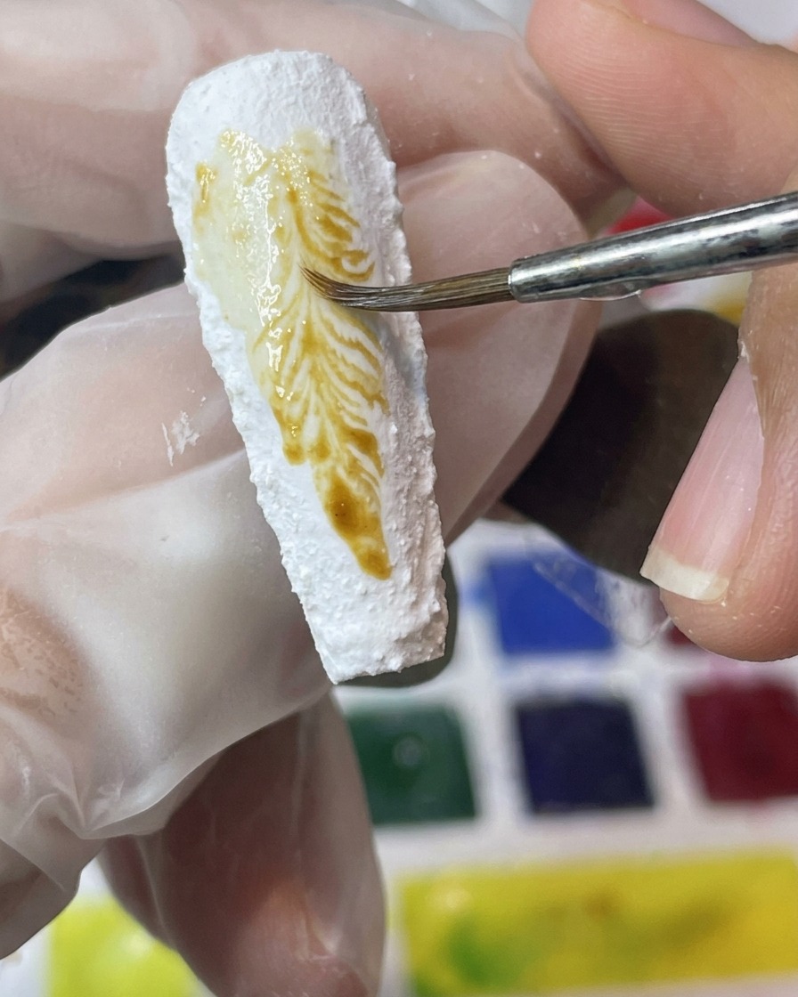 Un pincel aplicando pigmentos amarillos y marrones sobre una superficie de uña blanca texturizada.