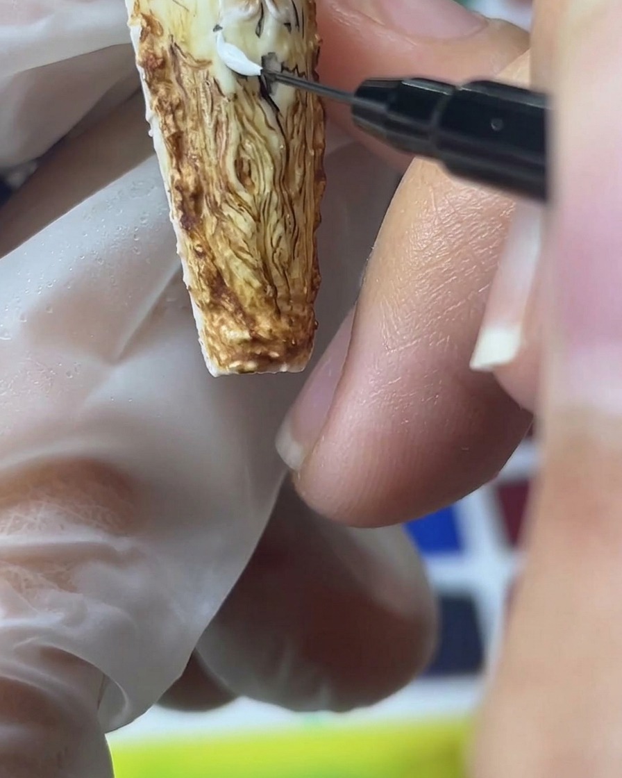 Primer plano de un pincel de detalle fino aplicando gel de arena blanco para crear pétalos de rosa dimensionales y texturizados sobre un fondo marrón.