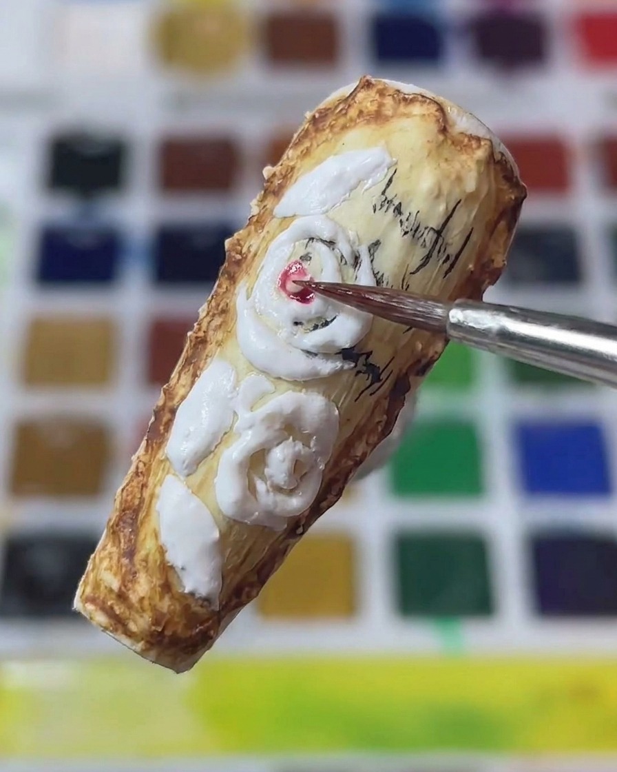 Primer plano de un pincel de arte de uñas fino aplicando pigmento rojo al centro de las rosas blancas esculpidas en 3D sobre una uña de estilo vintage.