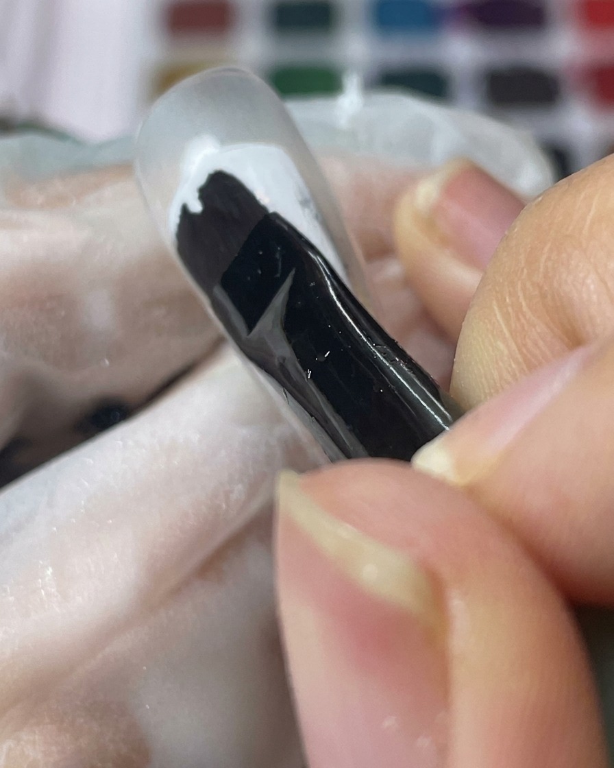 Un pincel de gel negro aplicando una capa suave de gel blanco sobre un tip de uña transparente.
