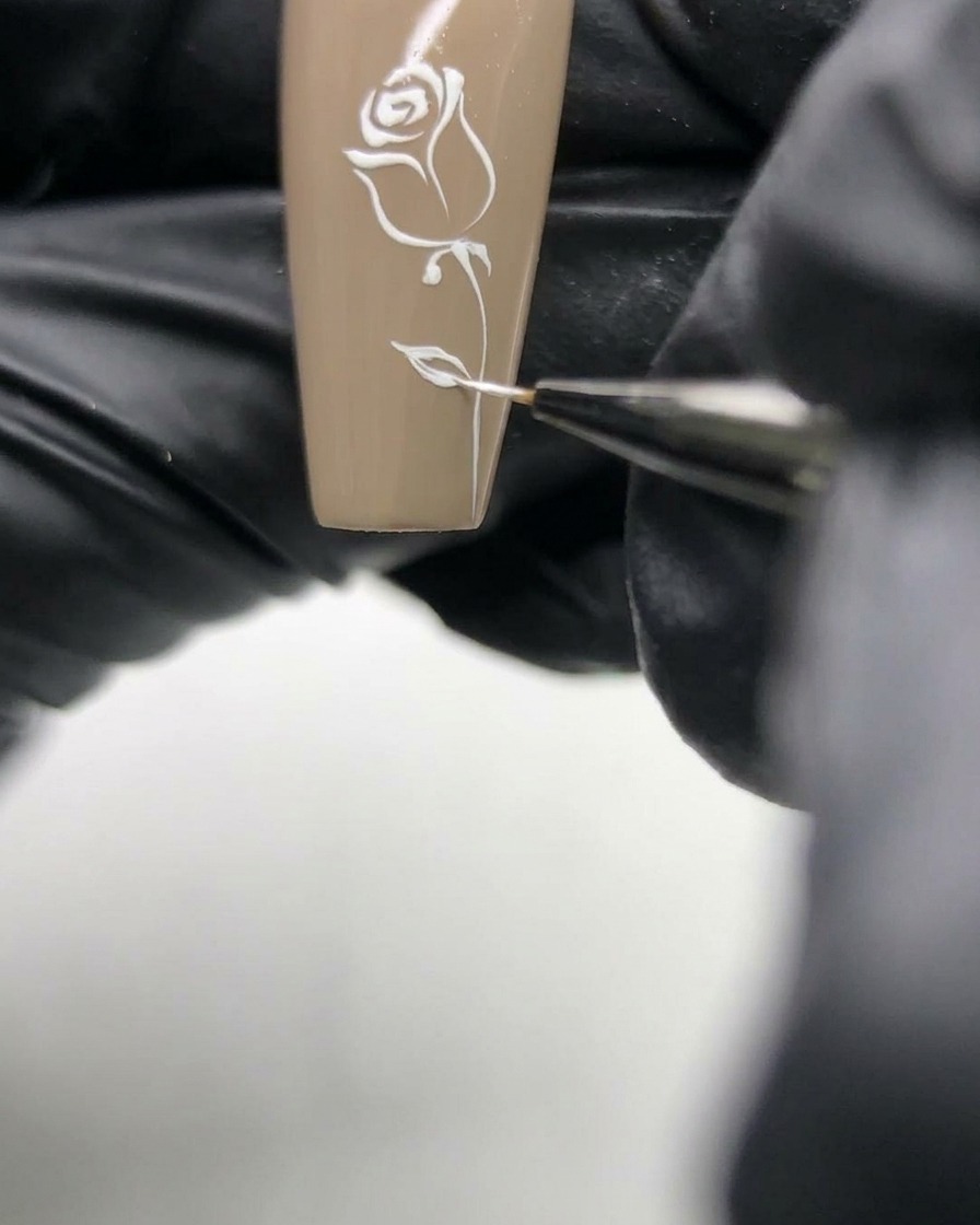 Macro de una manicurista refinando las delicadas líneas blancas de un diseño de rosa sobre una uña de base neutra.