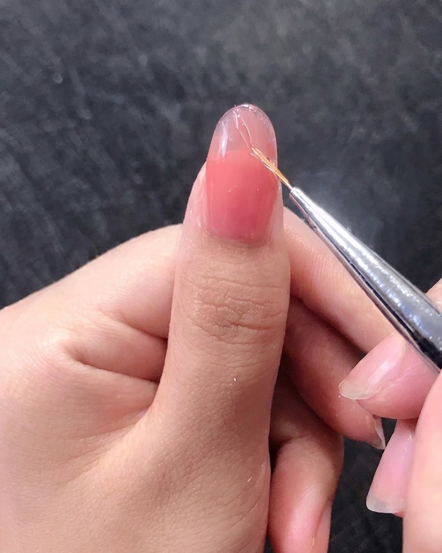 Primer plano de un pincel dibujando finas líneas doradas sobre una uña de gel rosa.
