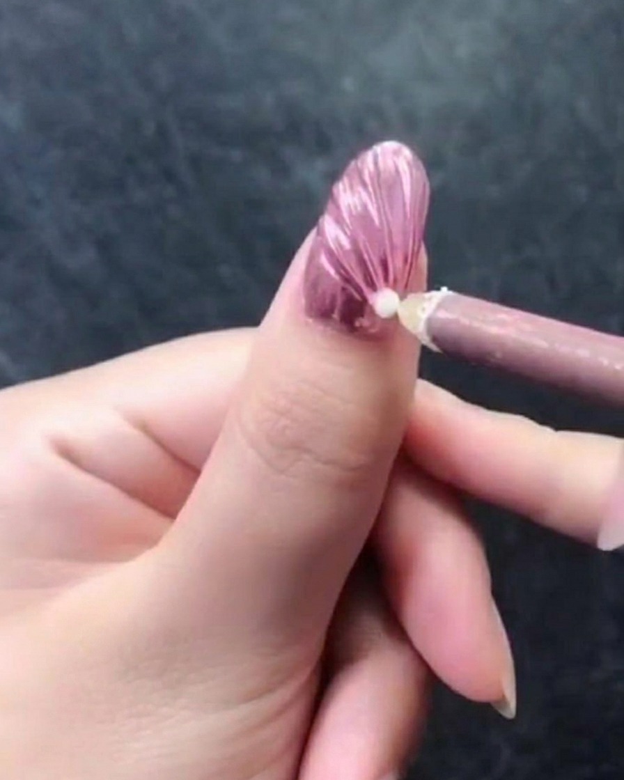 Primer plano de una mano con una uña metálica rosa texturizada, mostrando un lápiz de cera rosa colocando una perla blanca cerca de la zona de la cutícula.