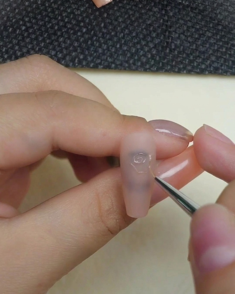 Un primer plano de una mano usando un pincel delineador fino para aplicar gel de escultura 3D transparente, dibujando el remolino central preciso de una rosa sobre una uña nude mate.