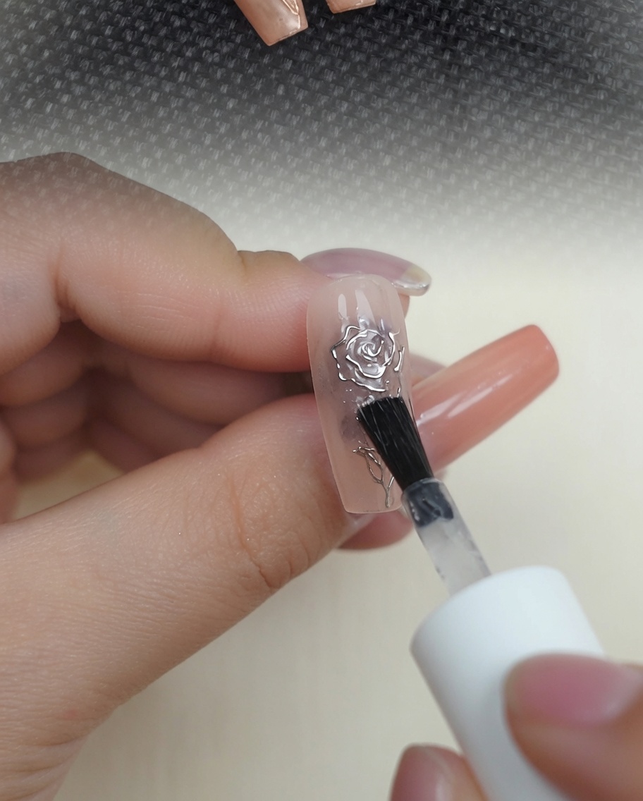 Un pincel aplicando una capa superior transparente y brillante sobre un diseño de rosa 3D cromada metálica en una uña nude.