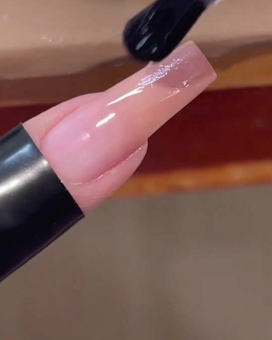 Un pincel de uñas aplicando una capa superior transparente y brillante sobre la uña con degradado rosa terminada.