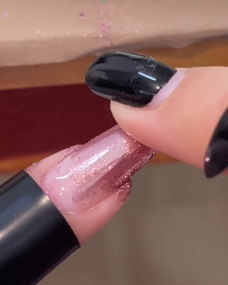 La yema de un dedo frotando polvo de cromo oro rosa sobre una uña terminada y brillante.