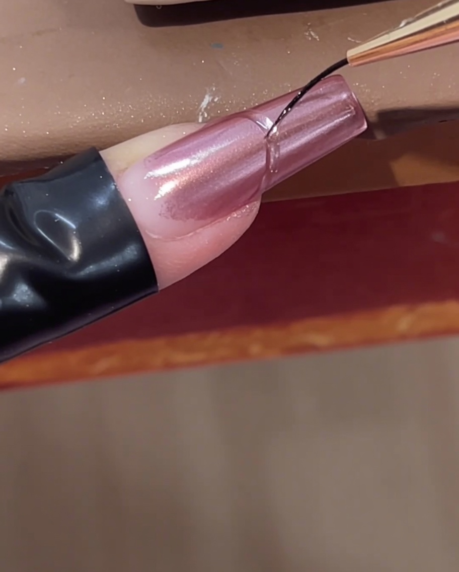 Un pincel delineador fino añadiendo una línea de gel constructor transparente curva cerca de la cutícula de una uña cromo rosa.