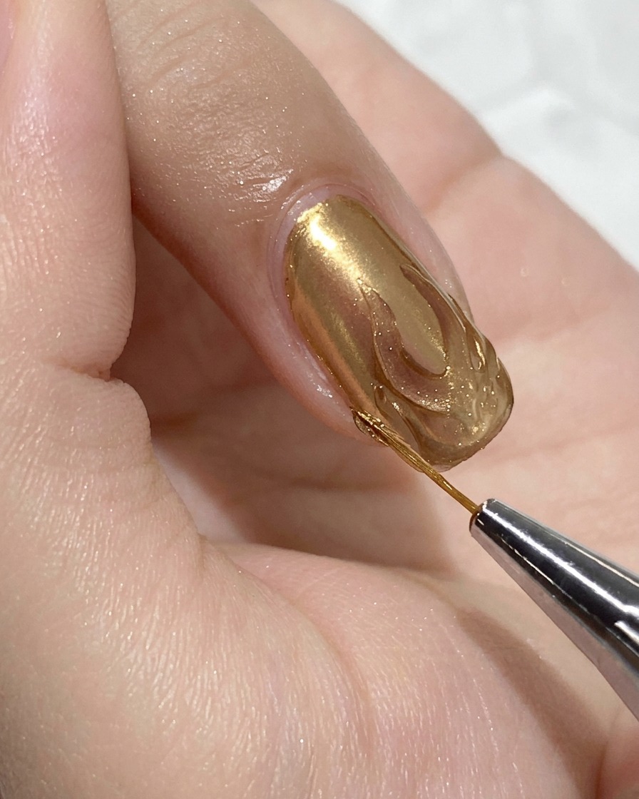 Primer plano de un pincel de detalle fino aplicando top coat de gel transparente dentro del contorno de una llama dorada sobre una uña rosa translúcida.