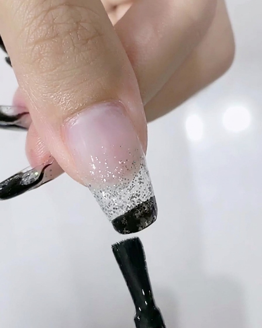 Un pincel aplicando esmalte de gel negro en el borde libre de una uña con degradado plateado.