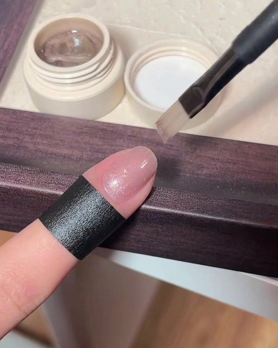 Vista en primer plano de un pincel aplicando un esmalte de gel cat eye rosa traslúcido y brillante sobre una uña natural.