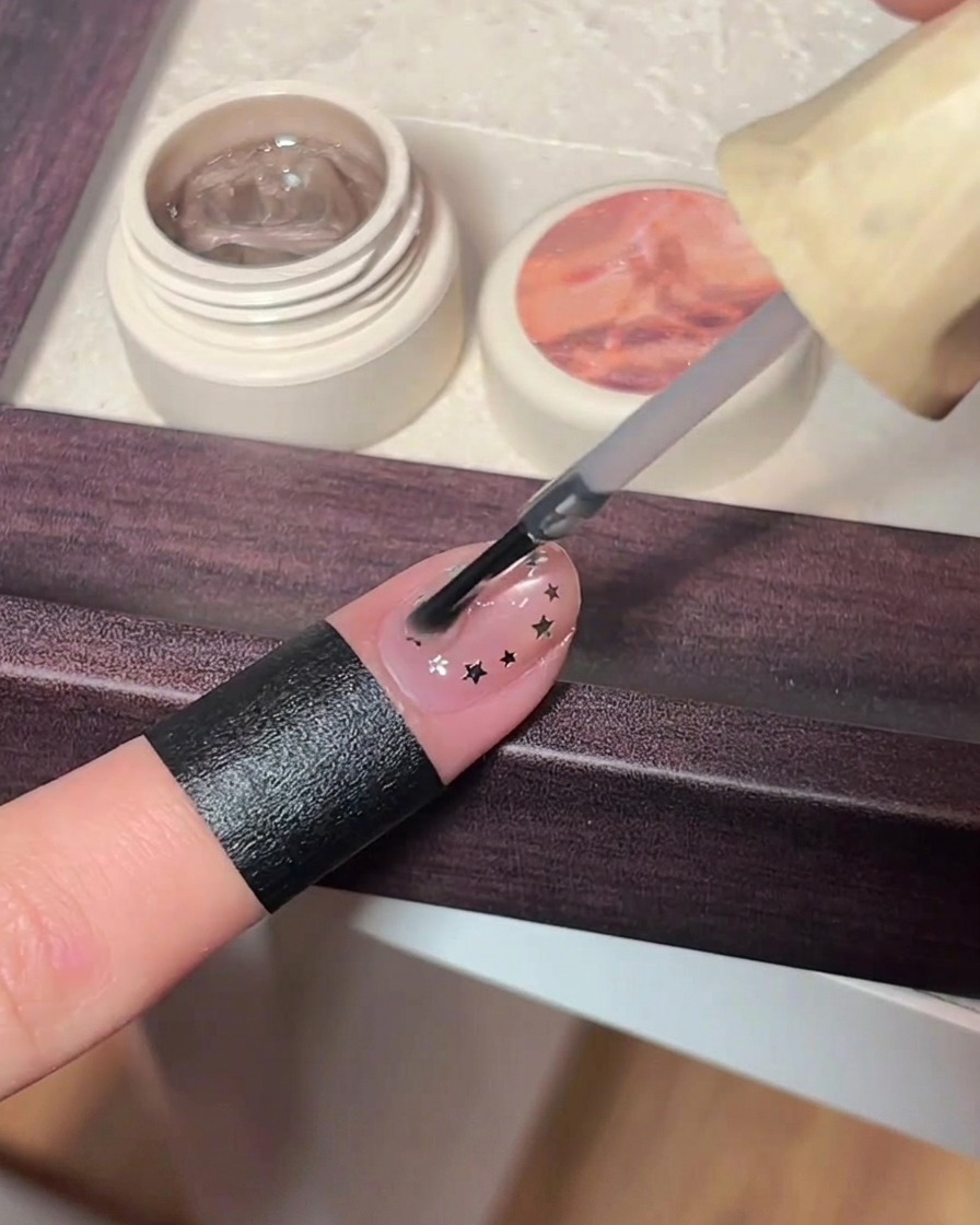 Un pincel aplicando una capa generosa de top coat transparente sobre una uña decorada con estrellas metálicas.