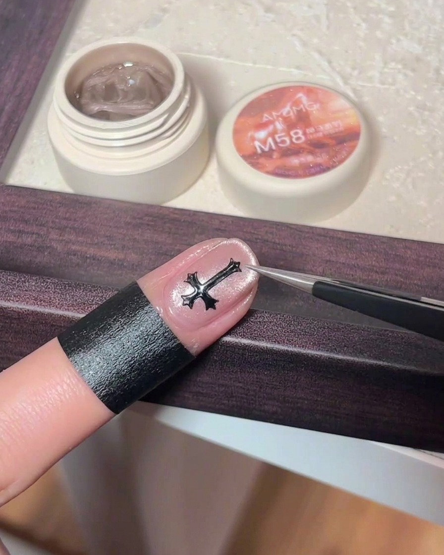 Pinzas colocando un dije de cruz metálico oscuro sobre el centro de una uña con gel cat eye rosa.