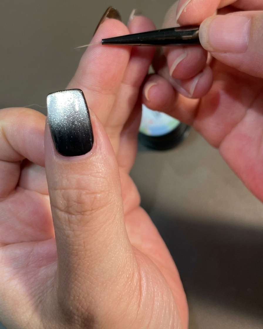 Primer plano de una uña siendo decorada con un pincel delineador fino creando líneas curvas similares a llamas sobre una base de gel magnético negro.