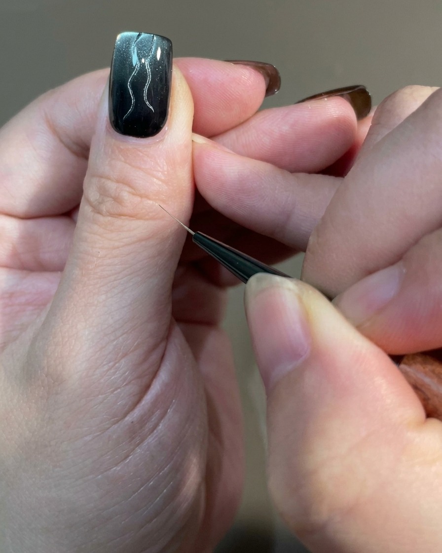 Primer plano de una uña mostrando múltiples líneas curvas de diferentes longitudes siendo dibujadas con un pincel delineador sobre una base de gel magnético.