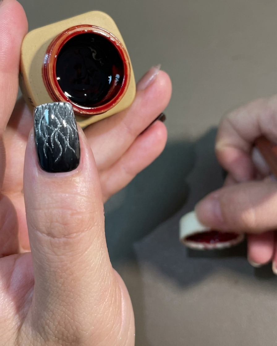 Un pincel de arte de uñas aplicando un gel rojo oscuro transparente sobre un diseño de llama ojo de gato blanco y negro curado.