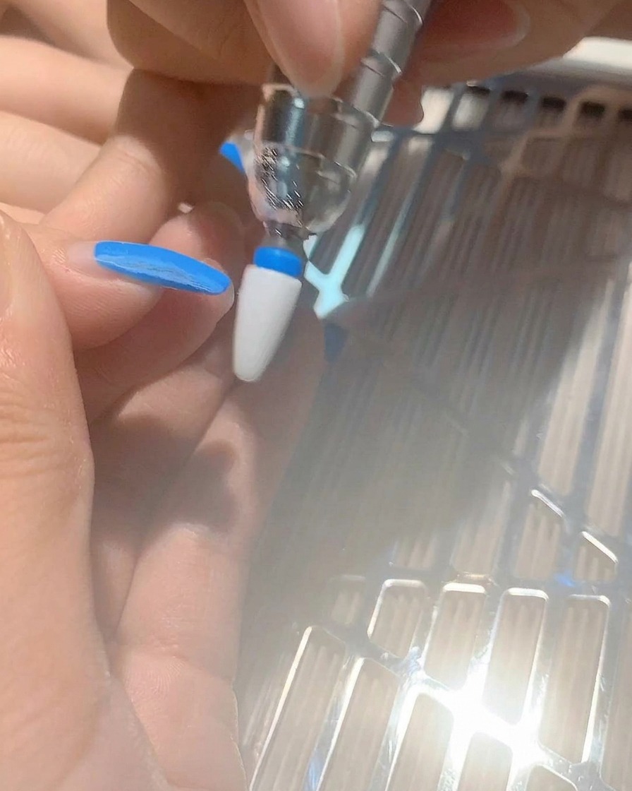 Primer plano de una fresa de torno retirando esmalte de gel azul de una uña.