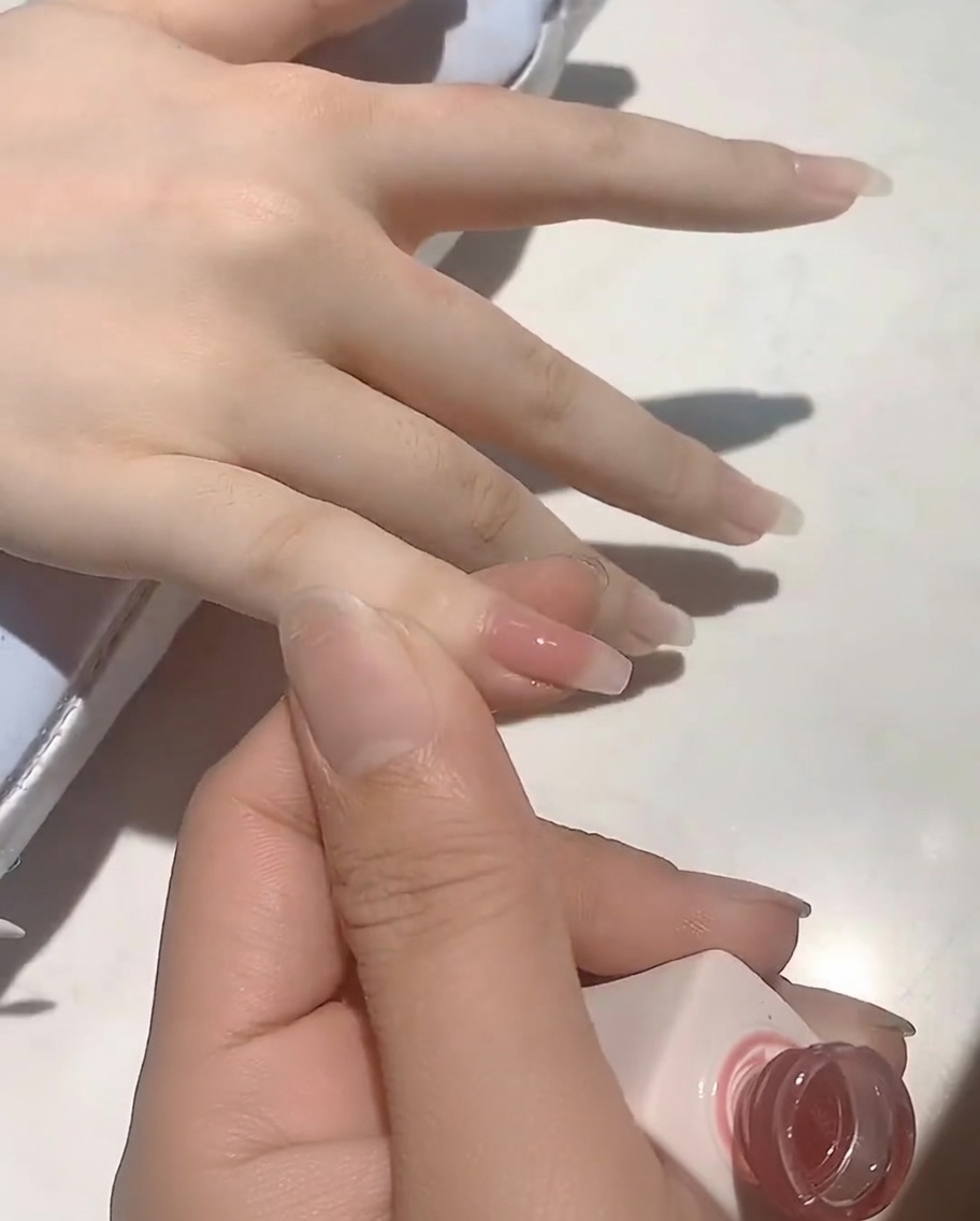 Un pincel aplicando un suave esmalte de gel rosa nude sobre una uña natural.