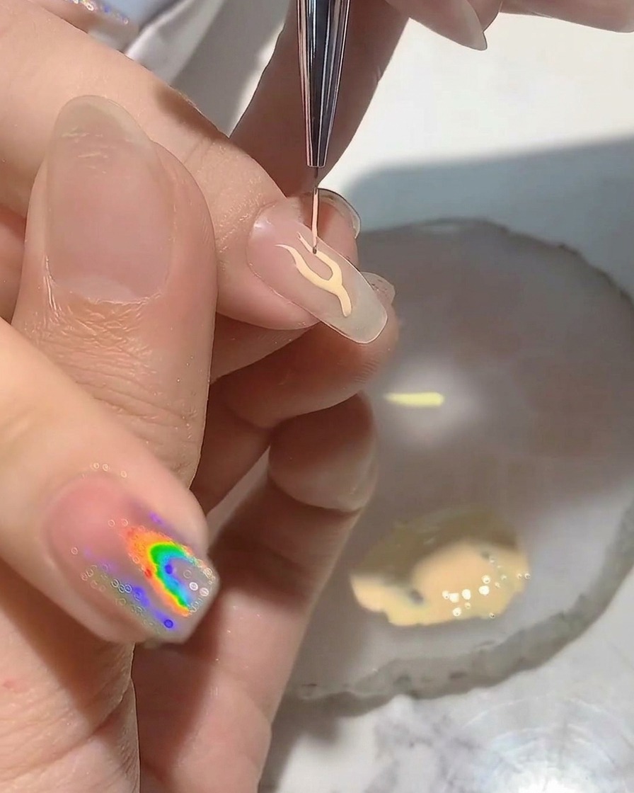 Una artista de uñas usando un pincel delineador fino para pintar el contorno de una llama delicada en un tip transparente.