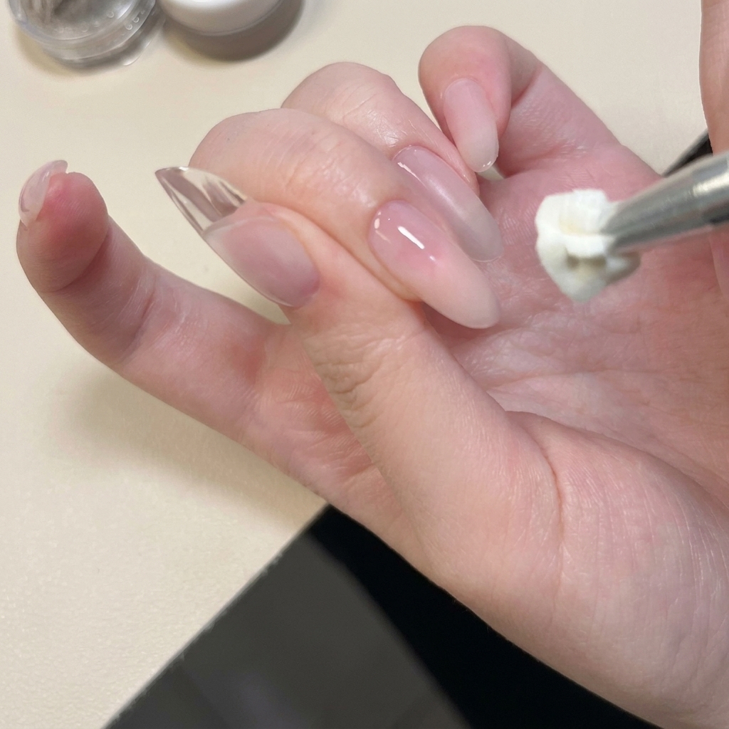 Primer plano de una herramienta metálica aplicando y presionando gel sólido blanco sobre la punta de una uña natural.