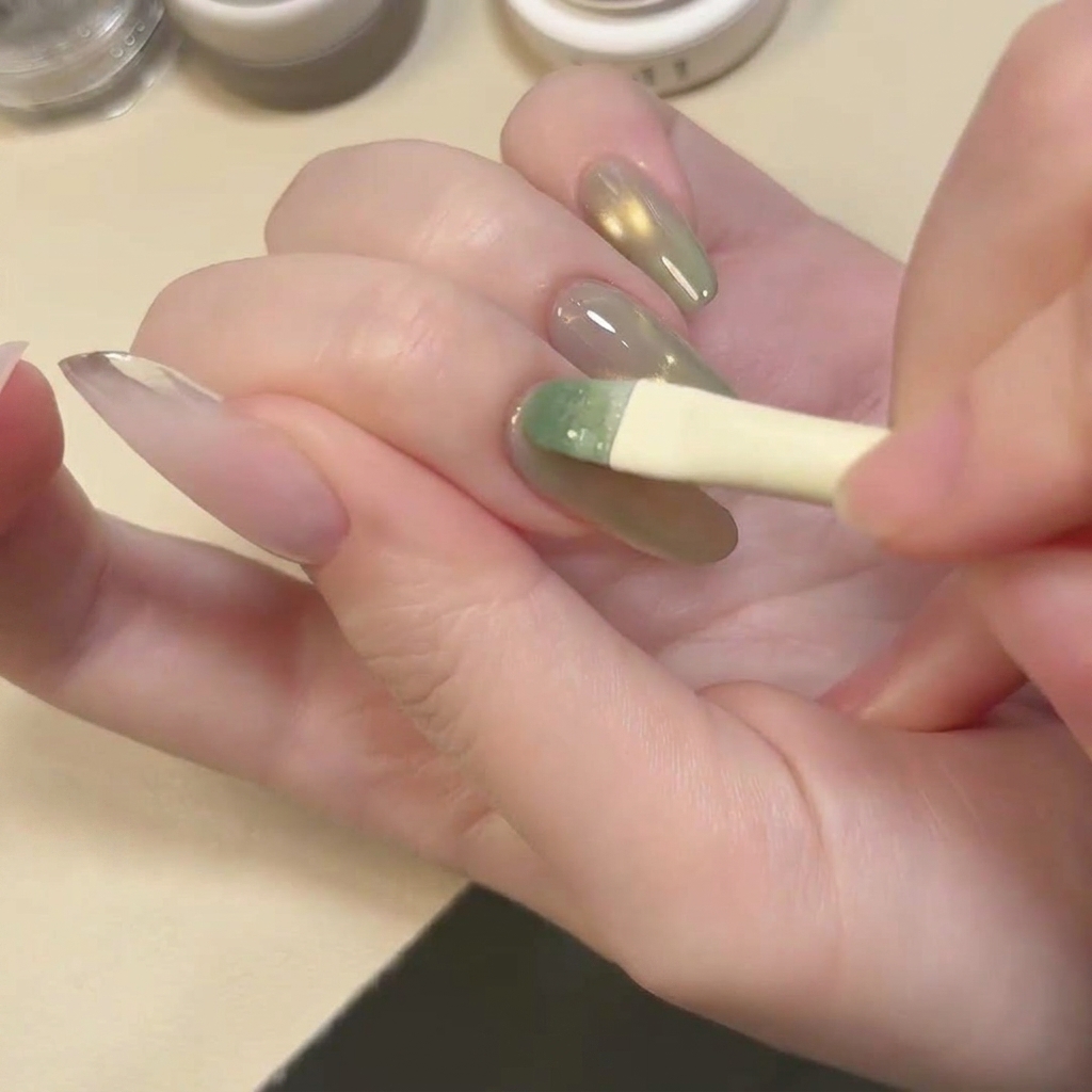 Un pincel aplicando una capa de esmalte de gel cat eye traslúcido en color verde lago sobre la uña.