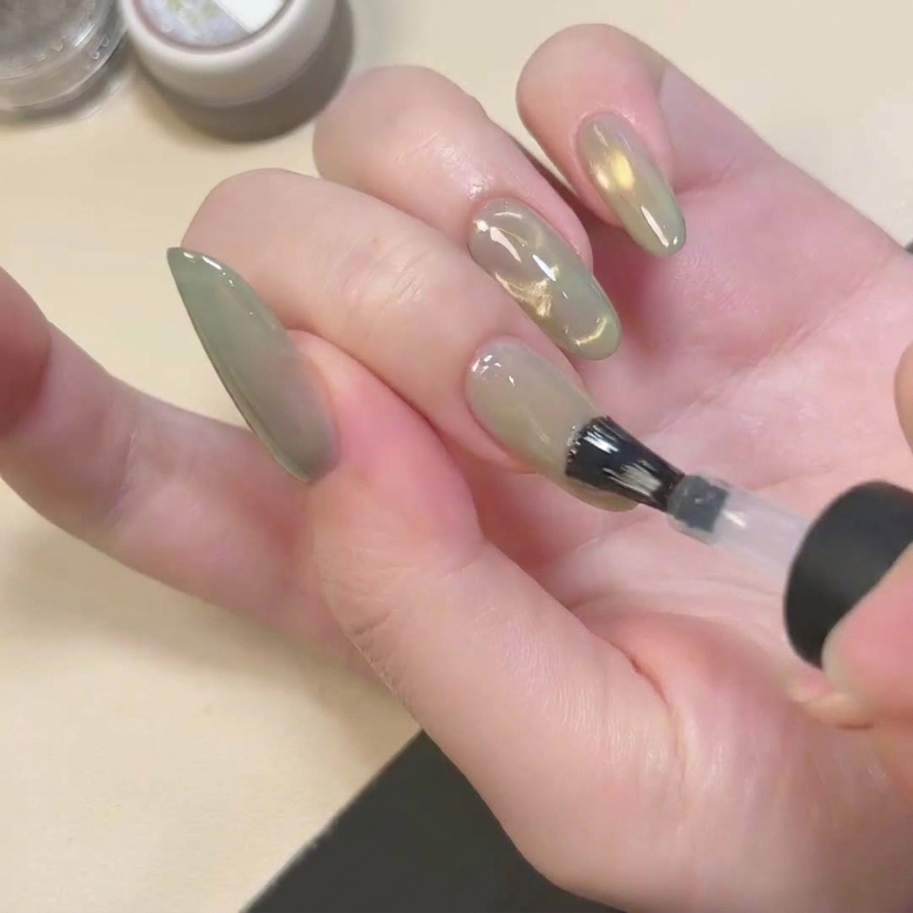 Un pincel aplicando un top coat transparente con acabado mate sobre una uña con efecto cat eye verde.