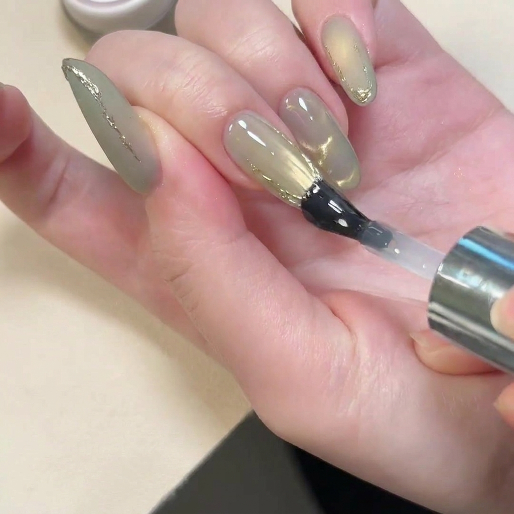 Un pincel aplicando un top coat brillante sobre una uña que tiene un acabado mate y detalles dorados en 3D.