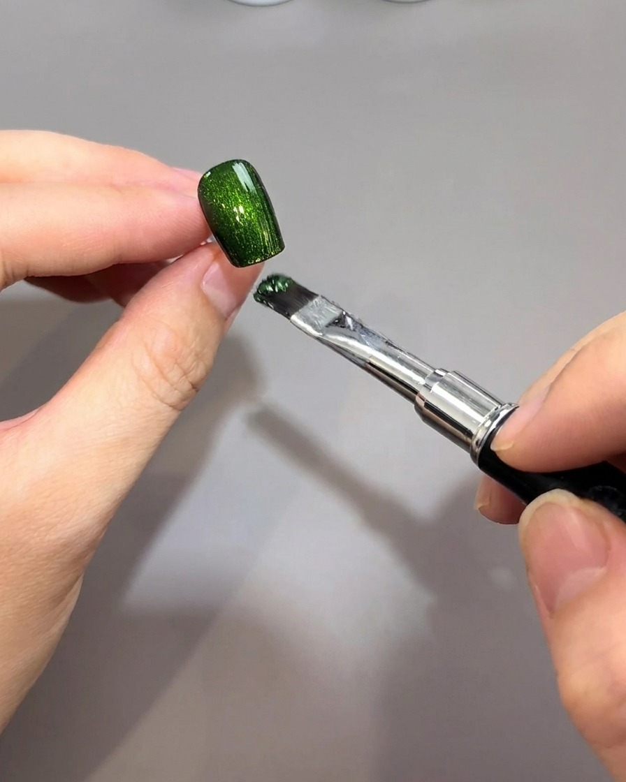 Un pincel aplicando un vibrante gel magnético verde esmeralda sobre una base de uña negra.