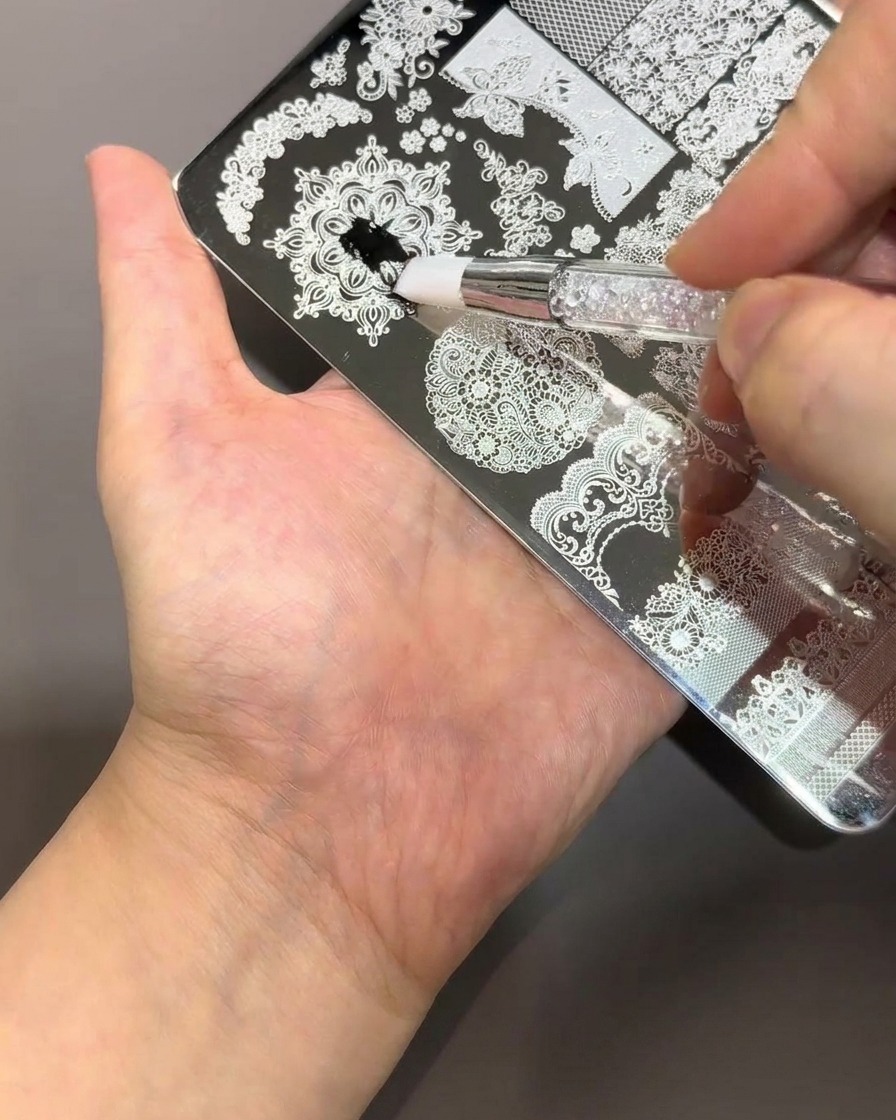 Un pincel aplicando esmalte negro espeso sobre una placa de metal plateado para stamping con intrincados patrones florales y de encaje.