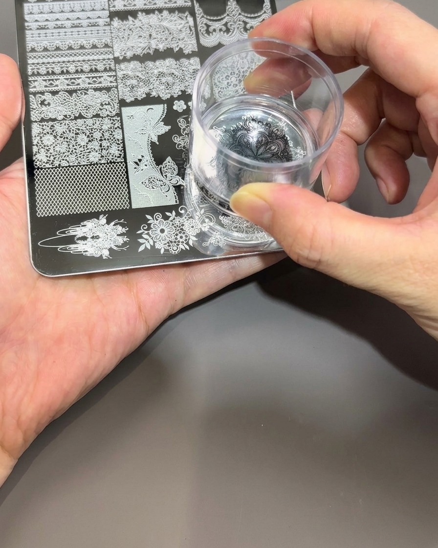 Una mano sosteniendo un estampador de uñas de silicona transparente, presionándolo suavemente sobre el diseño de esmalte negro en una placa de metal para stamping.