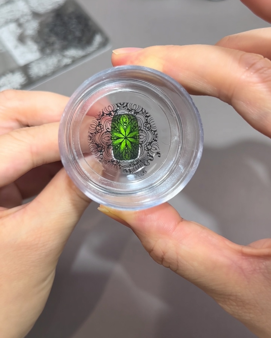 Mirando directamente hacia abajo a través de un estampador de silicona transparente, mostrando un intrincado patrón negro perfectamente alineado sobre una uña verde brillante con efecto ojo de gato.