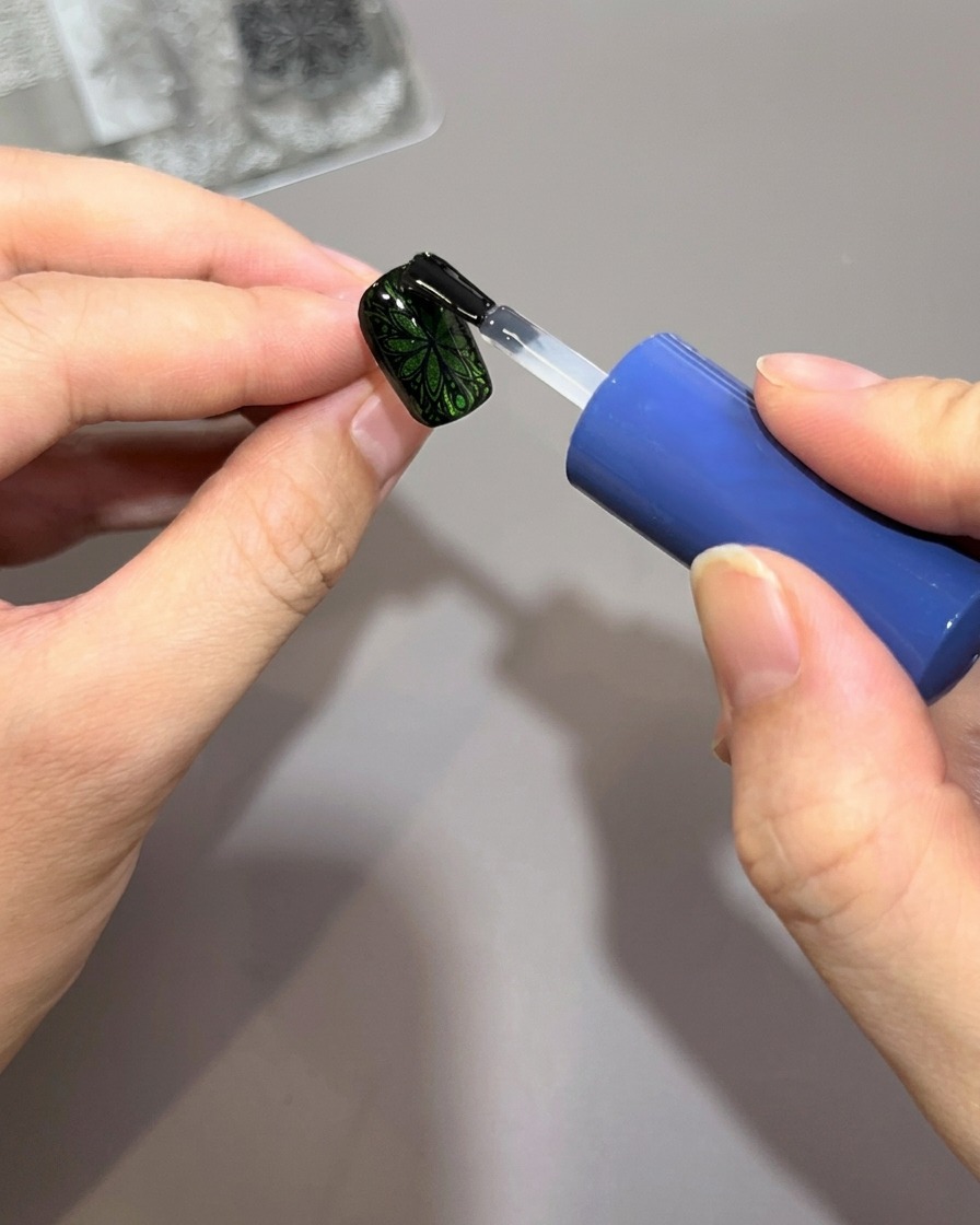 Un pincel de esmalte aplicando una capa gruesa de top coat transparente y brillante sobre una uña verde decorada con un intrincado patrón negro estampado.